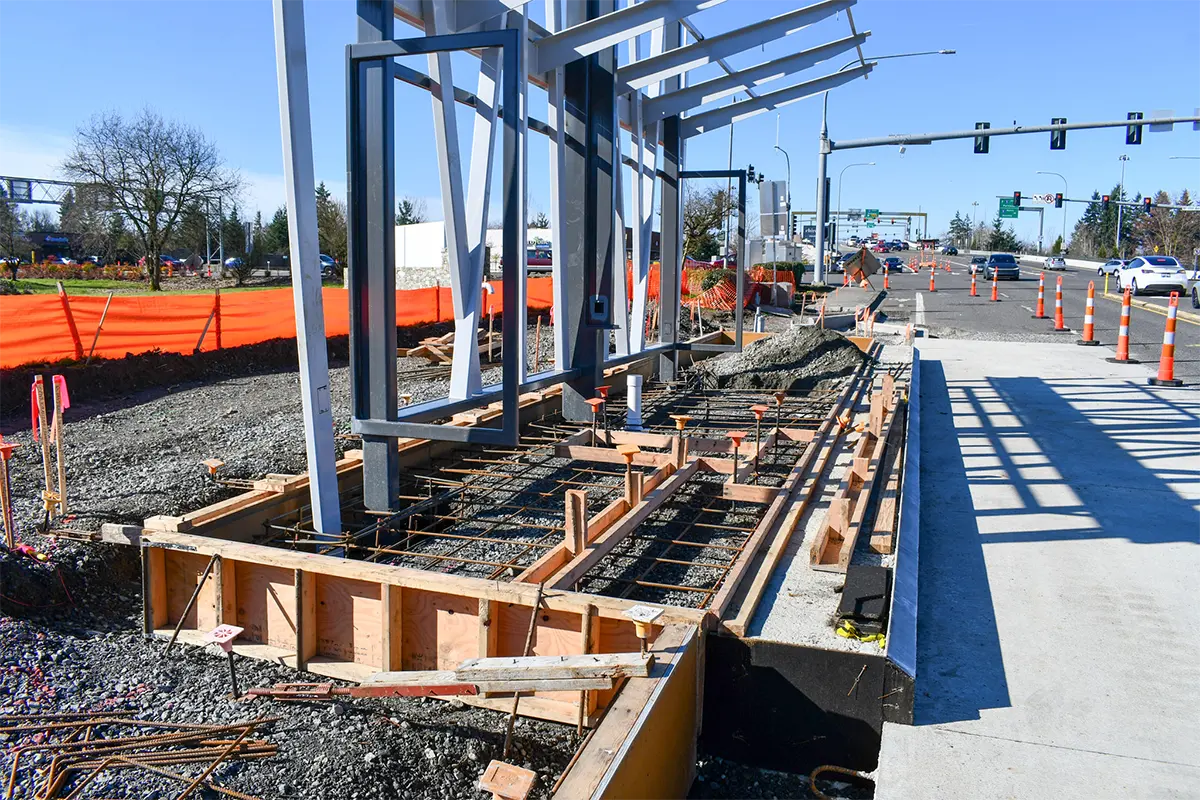 Concrete foundation work for a bus rapid transit station along Highway 99, with steel frame supports, rebar, and construction barriers beside an active roadway.