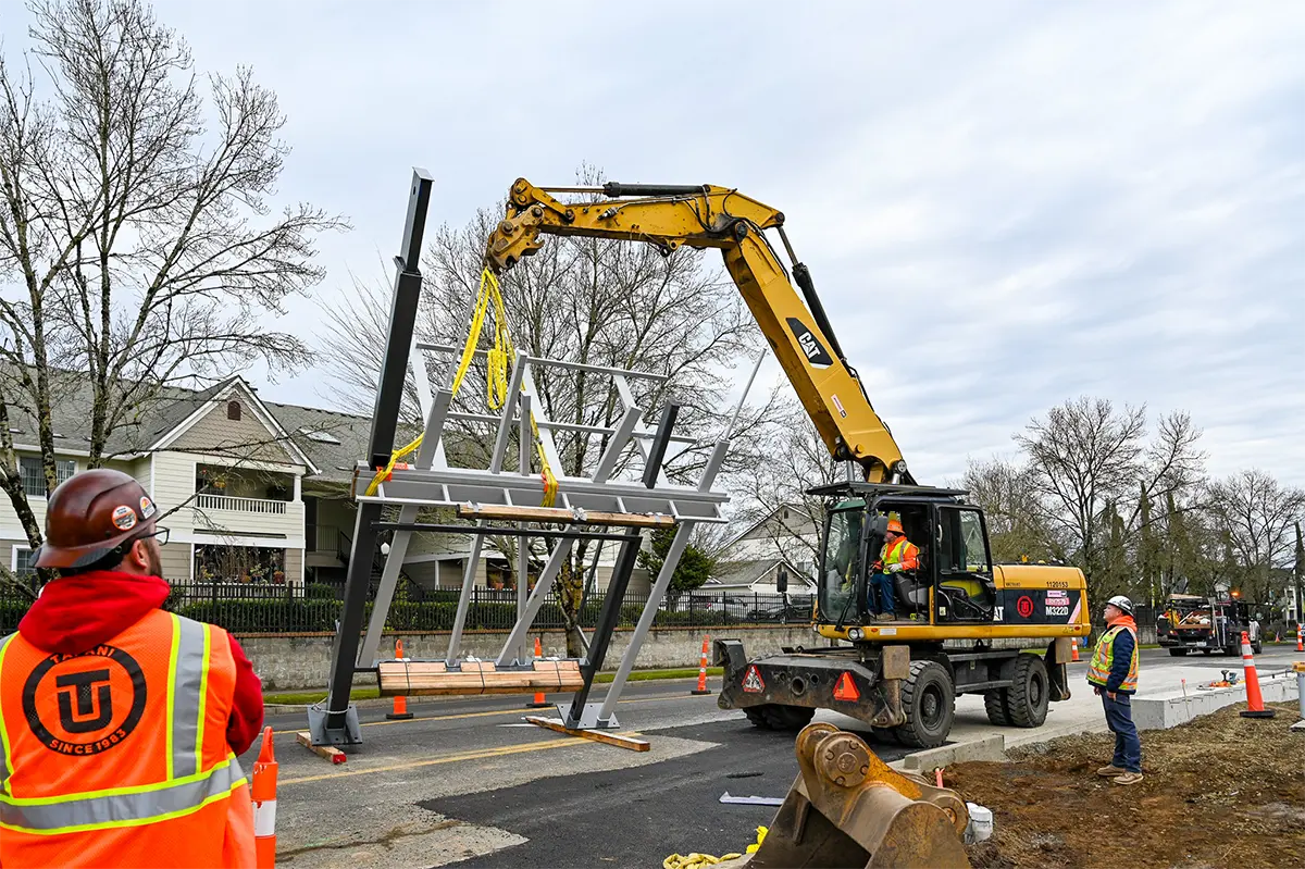 Excavator lifting and positioning a prefabricated steel shelter structure for a Highway 99 BRT station, with construction workers guiding the installation on a city street.